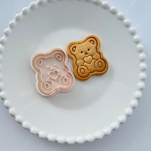 May include: A white ceramic plate with a scalloped edge features a cookie cutter and a cookie in the shape of a bear holding a heart. Both the cookie cutter and the cookie are light pink.