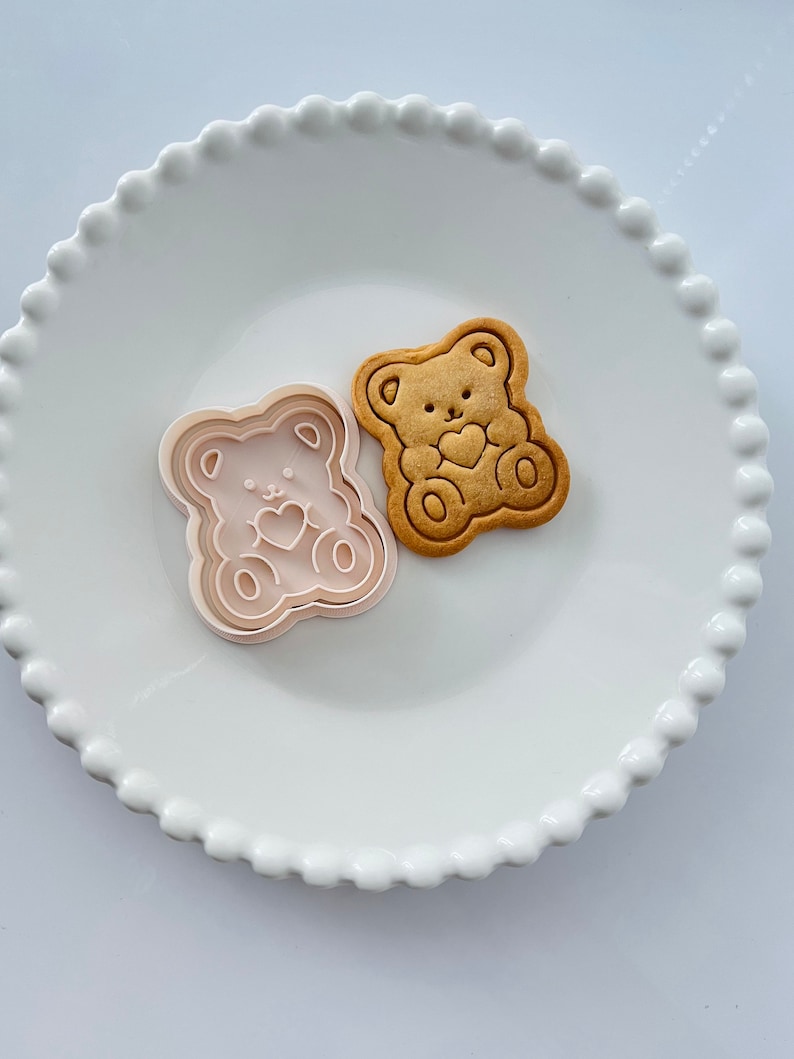 May include: A white ceramic plate with a scalloped edge features a cookie cutter and a cookie in the shape of a bear holding a heart. Both the cookie cutter and the cookie are light pink.