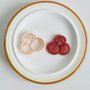 May include: A white plastic cookie cutter shaped like three cherries, next to a red cookie in the same shape.