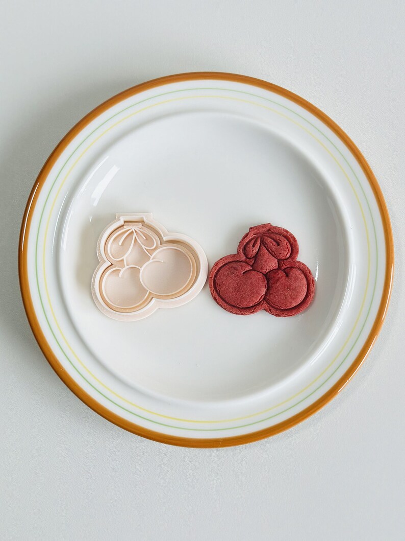 May include: A white plastic cookie cutter shaped like three cherries, next to a red cookie in the same shape.