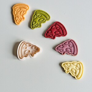 May include: Five different colored cookie cutters in the shape of a pizza slice. The cookie cutters are arranged in a semi-circle on a light gray surface. The cookie cutters are yellow, green, red, purple, and light yellow.