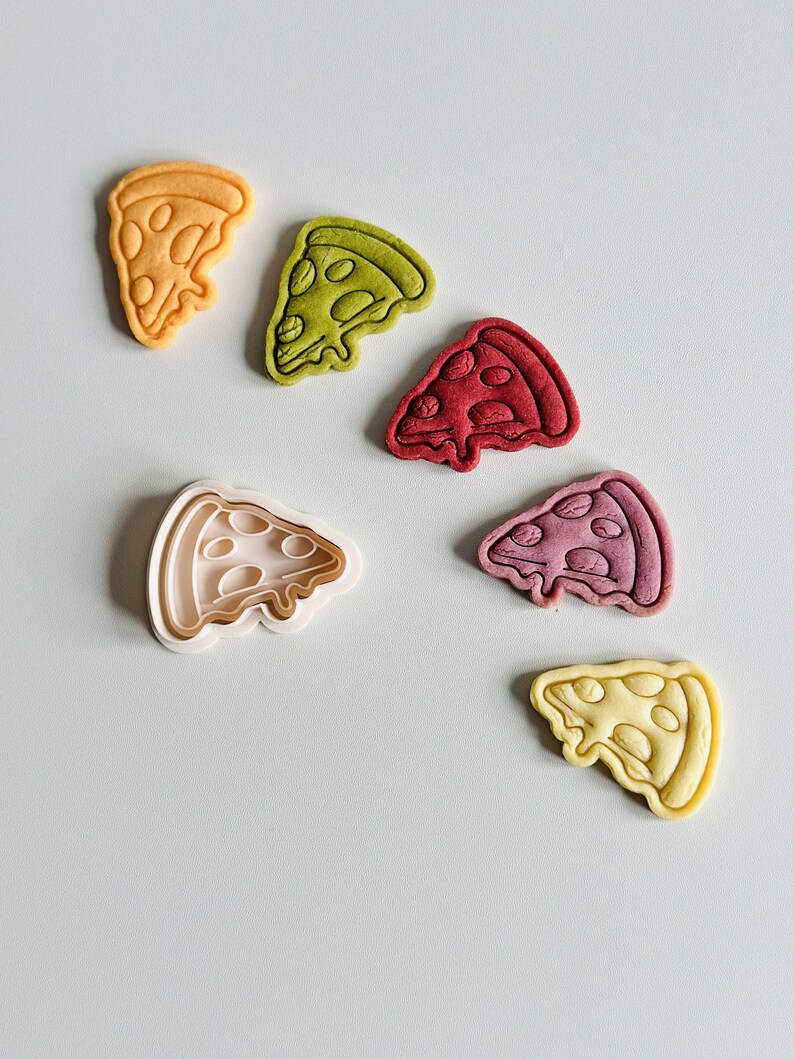 May include: Five different colored cookie cutters in the shape of a pizza slice. The cookie cutters are arranged in a semi-circle on a light gray surface. The cookie cutters are yellow, green, red, purple, and light yellow.