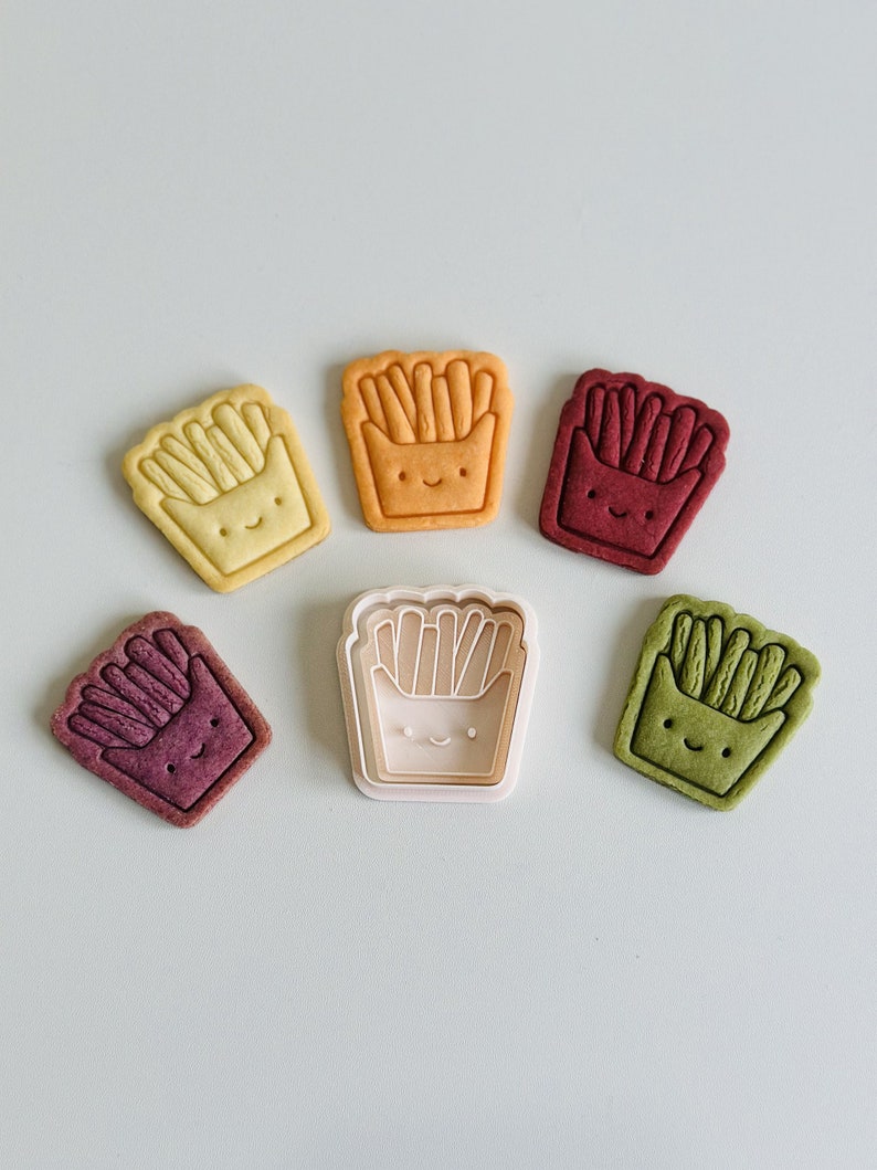 May include: Five cookie cutters in the shape of a cartoon french fry container with a smiling face. The cutters are in various colors: red, green, yellow, orange, and white. The white cutter is the only one that is not a cookie.