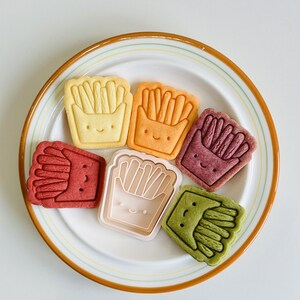May include: Six colorful cookies shaped like french fries in a box with a smiling face on a white plate with a brown rim.