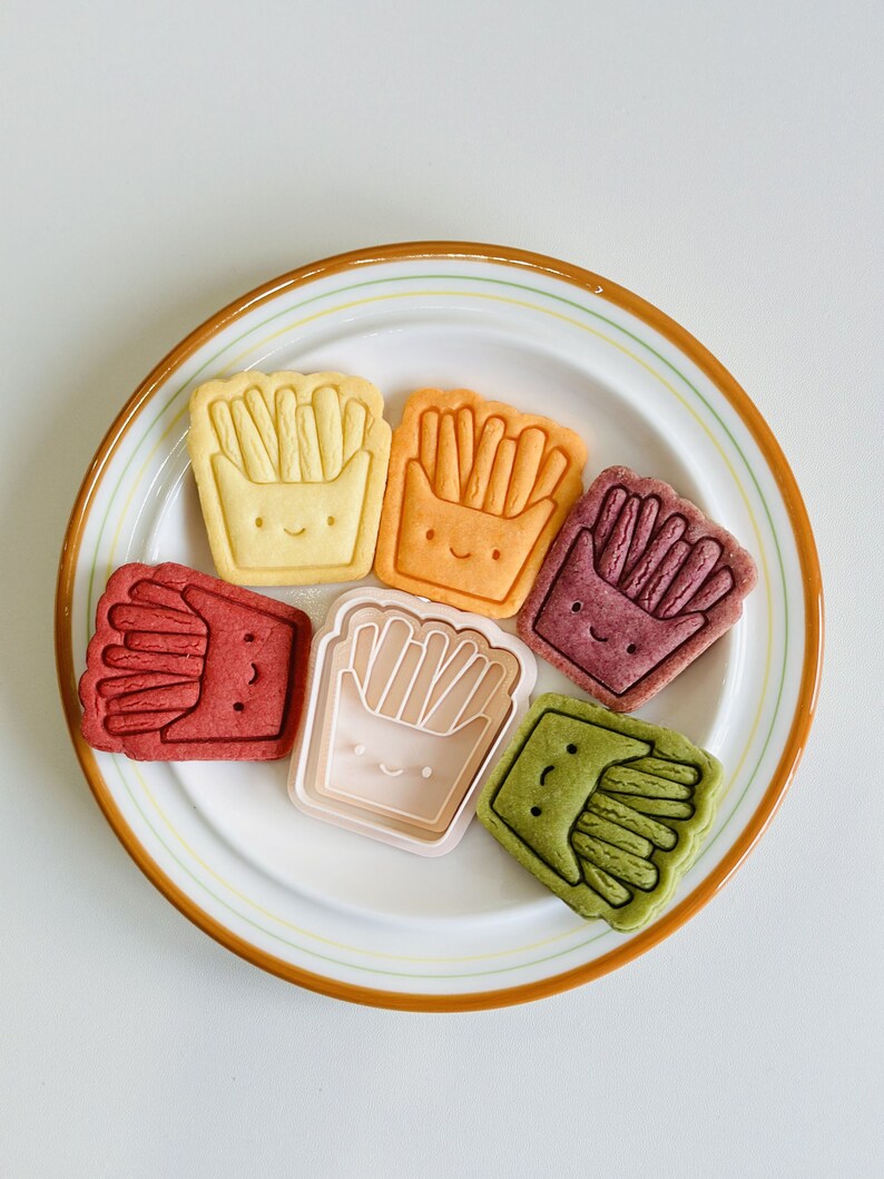 May include: Six colorful cookies shaped like french fries in a box with a smiling face on a white plate with a brown rim.