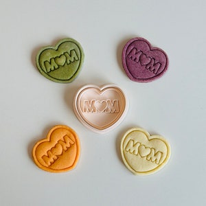 May include: Five heart-shaped cookies, four decorated with the word "MOM" and a heart, and one undecorated cookie cutter. The cookies are in shades of green, purple, orange, and yellow.