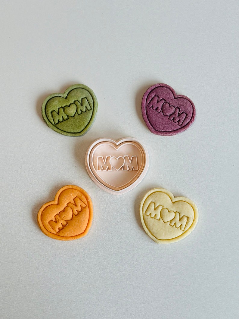 May include: Five heart-shaped cookies, four decorated with the word "MOM" and a heart, and one undecorated cookie cutter. The cookies are in shades of green, purple, orange, and yellow.