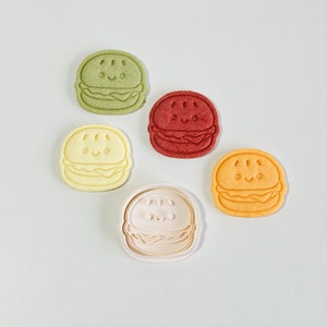 Puede incluir: Cinco cortadores de galletas de diferentes colores con forma de hamburguesa con una cara sonriente. Los cortadores son verdes, rojos, amarillos, naranjas y blancos.