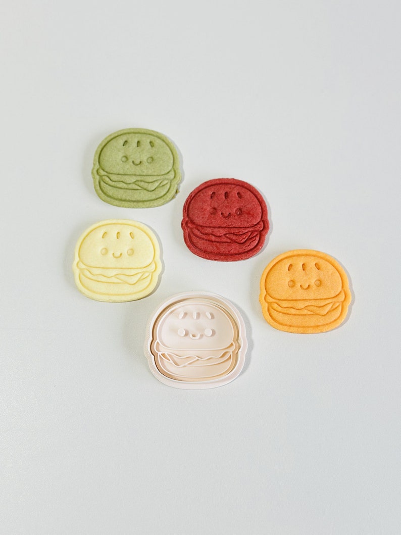 Puede incluir: Cinco cortadores de galletas de diferentes colores con forma de hamburguesa con una cara sonriente. Los cortadores son verdes, rojos, amarillos, naranjas y blancos.