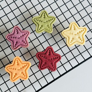 May include: Five star-shaped cookies on a black wire rack. The cookies are different colours: green, purple, red, orange, and yellow. Each cookie has a smiling face and a dotted pattern around the edge.