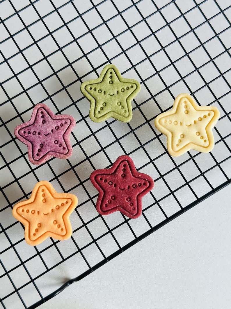 May include: Five star-shaped cookies on a black wire rack. The cookies are different colours: green, purple, red, orange, and yellow. Each cookie has a smiling face and a dotted pattern around the edge.