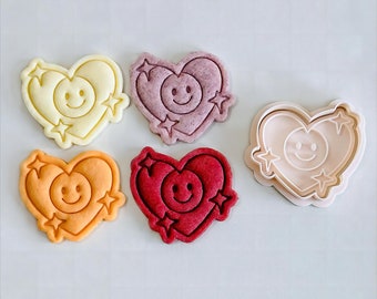 May include: Five heart-shaped cookie cutters with a smiley face and stars design. The cutters are in various colors: yellow, pink, red, and beige.