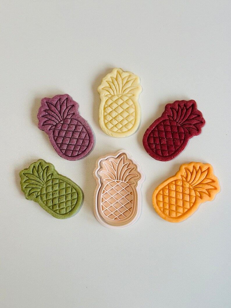 May include: Six pineapple-shaped cookies in various colors, including green, yellow, orange, red, and purple, with a white pineapple cookie cutter in the center.