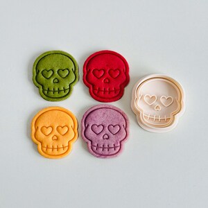 May include: A collection of colorful skull-shaped cookies and a cookie cutter. The cookies are in shades of green, red, orange, and purple, each with heart-shaped eyes. The cookie cutter is light pink.