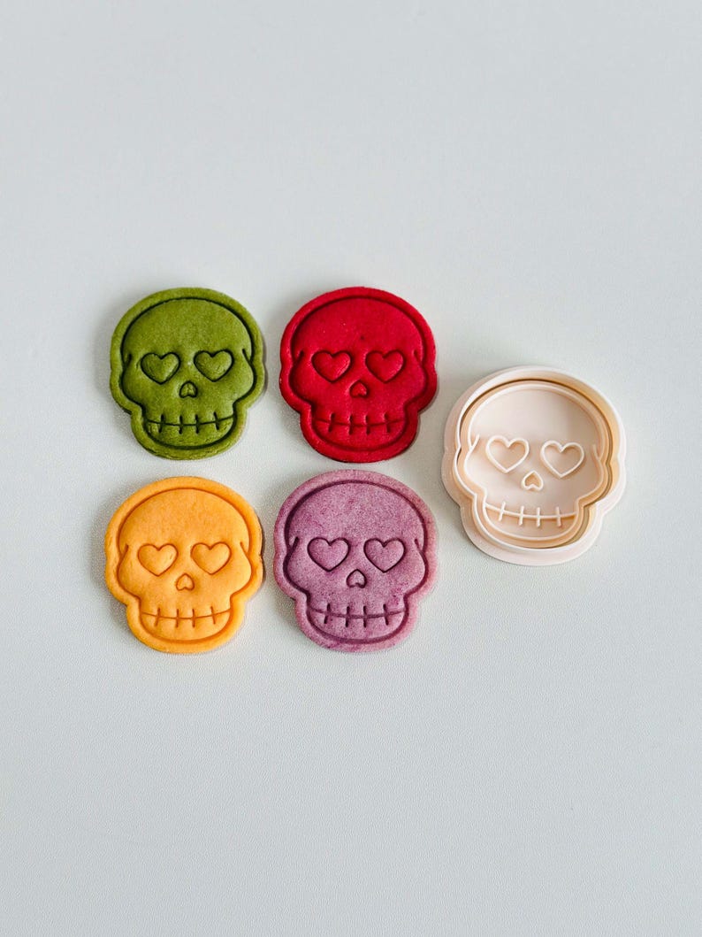May include: A collection of colorful skull-shaped cookies and a cookie cutter. The cookies are in shades of green, red, orange, and purple, each with heart-shaped eyes. The cookie cutter is light pink.
