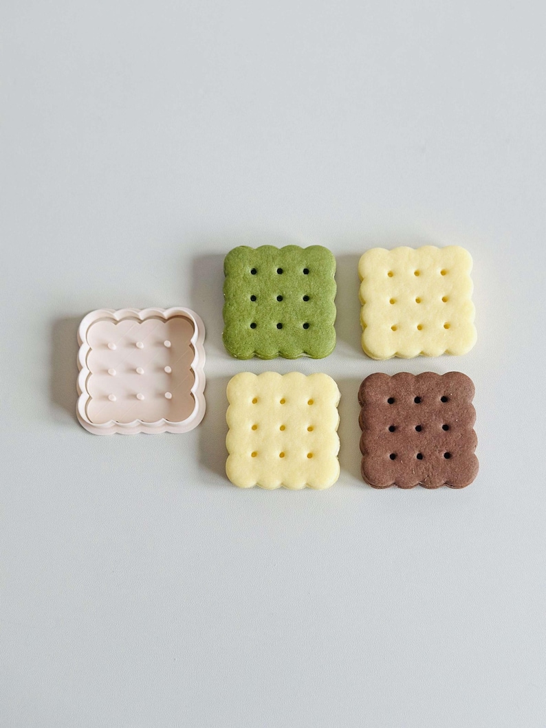 May include: Four square-shaped cookies, two yellow, one green, and one brown, with a plastic cookie cutter in the shape of a square with rounded edges.