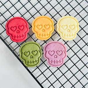 May include: Five colorful skull-shaped cookies with heart-shaped eyes and a cross-shaped mouth are arranged on a black wire rack. The cookies are in shades of red, orange, yellow, green, and purple. The cookies are a fun and festive treat.