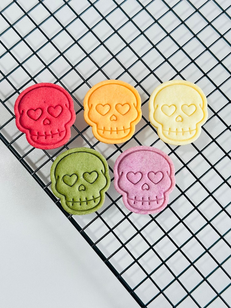 May include: Five colorful skull-shaped cookies with heart-shaped eyes and a cross-shaped mouth are arranged on a black wire rack. The cookies are in shades of red, orange, yellow, green, and purple. The cookies are a fun and festive treat.
