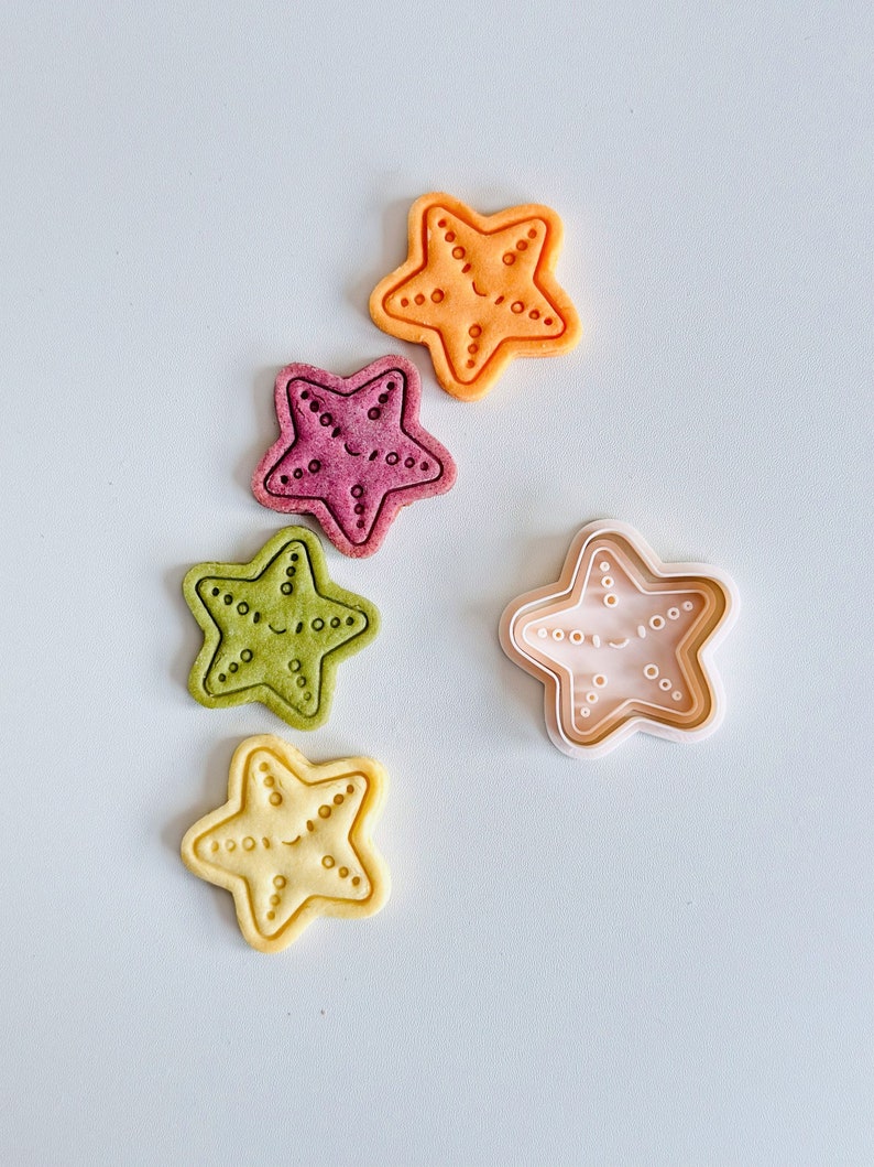 May include: Four starfish-shaped cookies in different colours: orange, purple, green, and yellow. The cookies have a smiling face and are arranged on a light blue surface. A white plastic cookie cutter in the shape of a starfish is also shown.