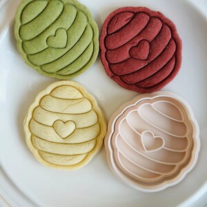 Pu&ograve; includere: Quattro biscotti a forma di favo con un cuore al centro. I biscotti sono verdi, rossi, gialli e beige. I biscotti sono su un piatto bianco.