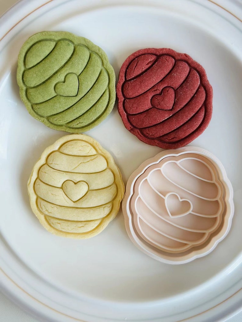Pu&ograve; includere: Quattro biscotti a forma di favo con un cuore al centro. I biscotti sono verdi, rossi, gialli e beige. I biscotti sono su un piatto bianco.
