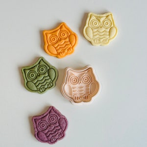 May include: Five owl-shaped cookie cutters, two are orange, one is green, one is purple, and one is white. The cutters are on a white surface.