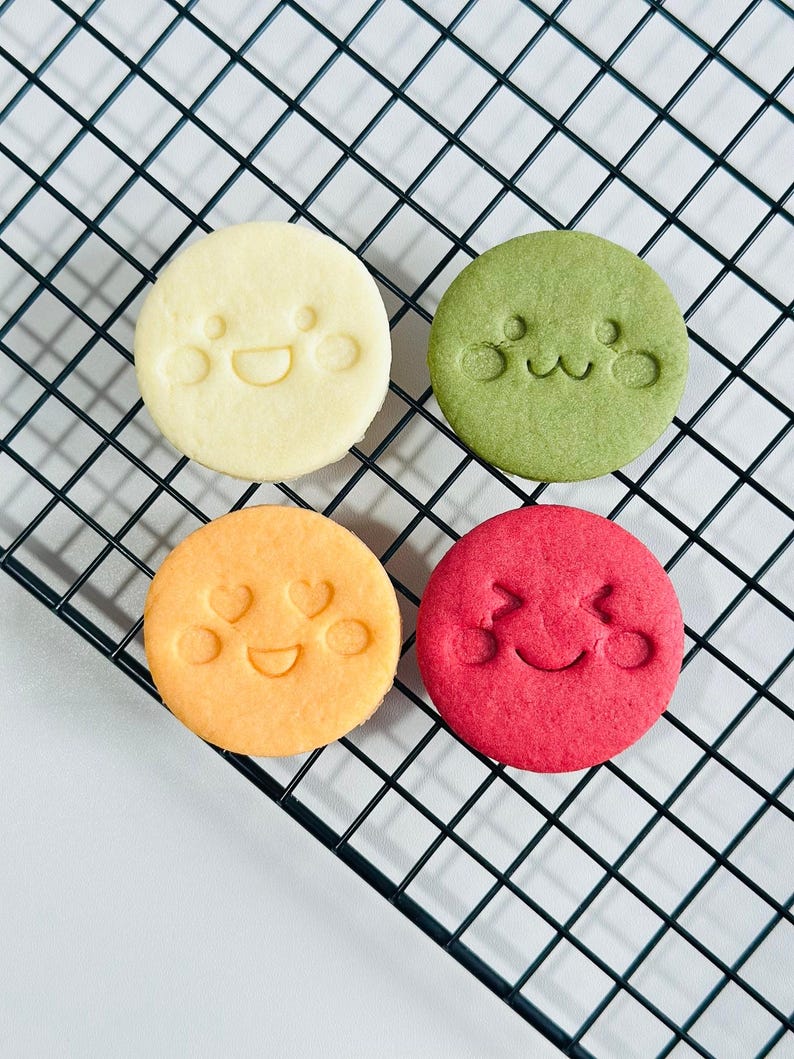 May include: Four round cookies with smiling faces on a black wire rack. The cookies are yellow, green, orange, and red. The cookies have a cute, playful design.