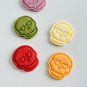 May include: An assortment of colorful skull-shaped cookies. Each cookie features heart-shaped eyes and a crossbones design. The cookies are in shades of purple, green, yellow, red, and orange. A fun and festive treat for Halloween or themed events.