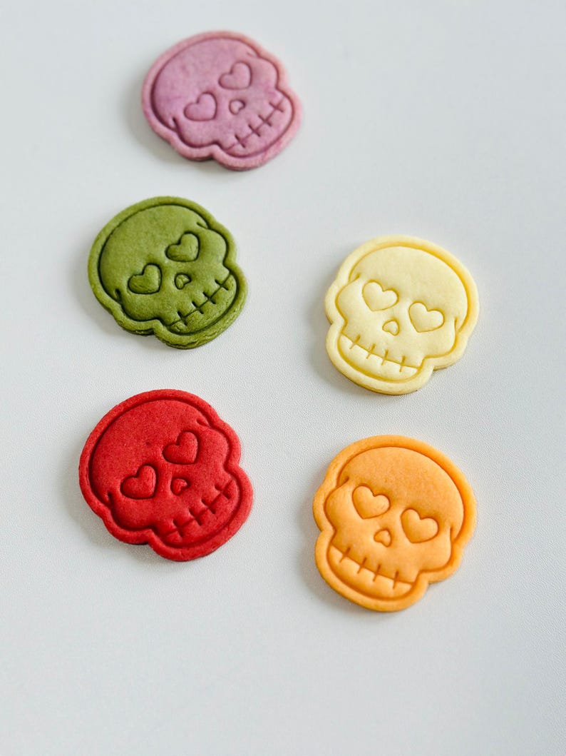 May include: An assortment of colorful skull-shaped cookies. Each cookie features heart-shaped eyes and a crossbones design. The cookies are in shades of purple, green, yellow, red, and orange. A fun and festive treat for Halloween or themed events.
