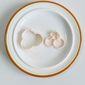 May include: Two plastic cookie cutters on a white plate with a brown rim. One cutter is shaped like a cloud and the other is shaped like a bunch of cherries.