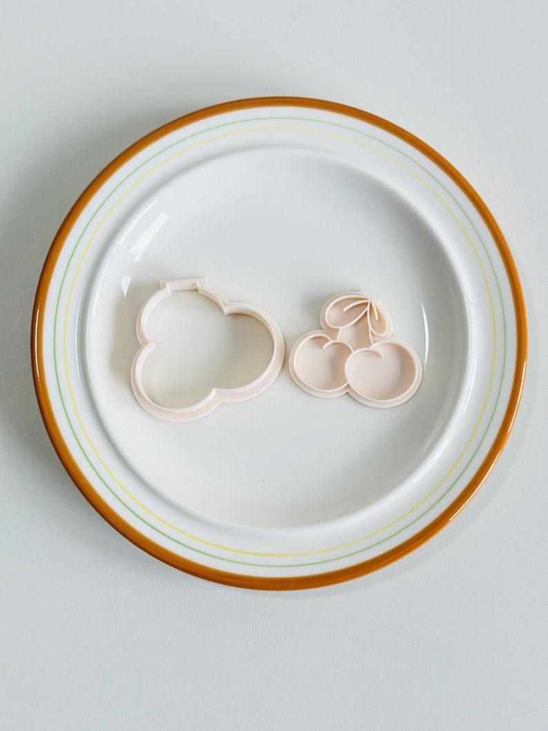 May include: Two plastic cookie cutters on a white plate with a brown rim. One cutter is shaped like a cloud and the other is shaped like a bunch of cherries.