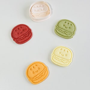 Puede incluir: Cinco cortadores de galletas con forma de hamburguesa con una cara sonriente. Los cortadores son de varios colores: blanco, rojo, verde, amarillo y naranja.