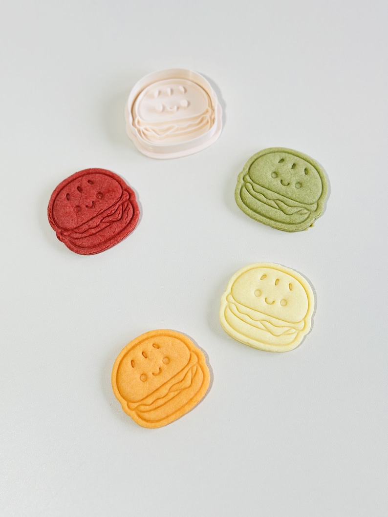 Puede incluir: Cinco cortadores de galletas con forma de hamburguesa con una cara sonriente. Los cortadores son de varios colores: blanco, rojo, verde, amarillo y naranja.