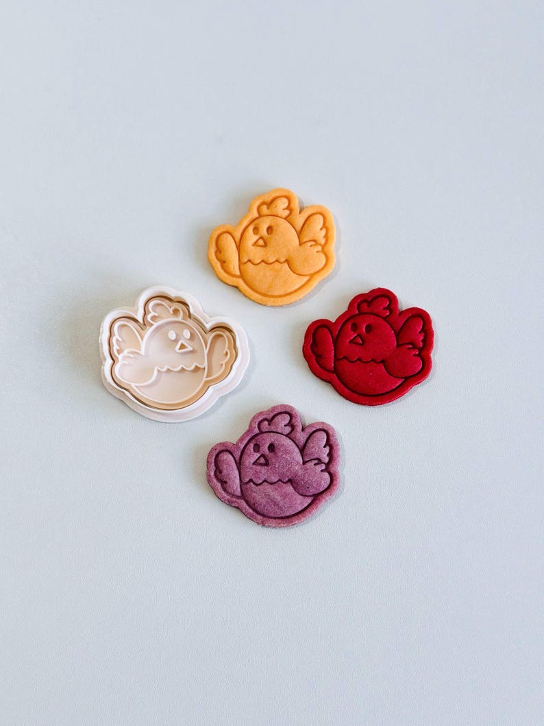 Op de afbeelding: Vier koekjessnijders en koekjes in de vorm van kuikens in verschillende kleuren: beige, oranje, rood en paars. De koekjes hebben gedetailleerde kenmerken, waaronder ogen, snavels en vleugels. De snijders zijn beige.