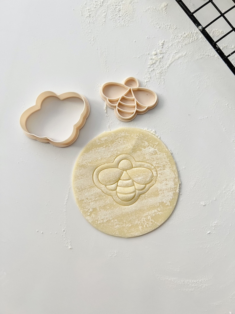 May include: Two cookie cutters, one shaped like a cloud and the other shaped like a bee, and a round piece of dough with a bee imprint.