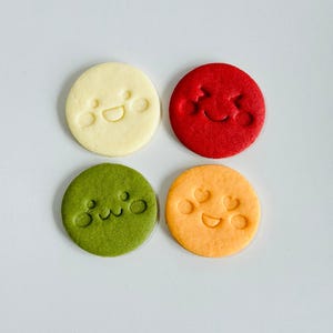 May include: Four round cookies in a square arrangement, each with a different color and a smiling face design. Colors include cream, red, green, and orange. The faces are created with simple, embossed lines.
