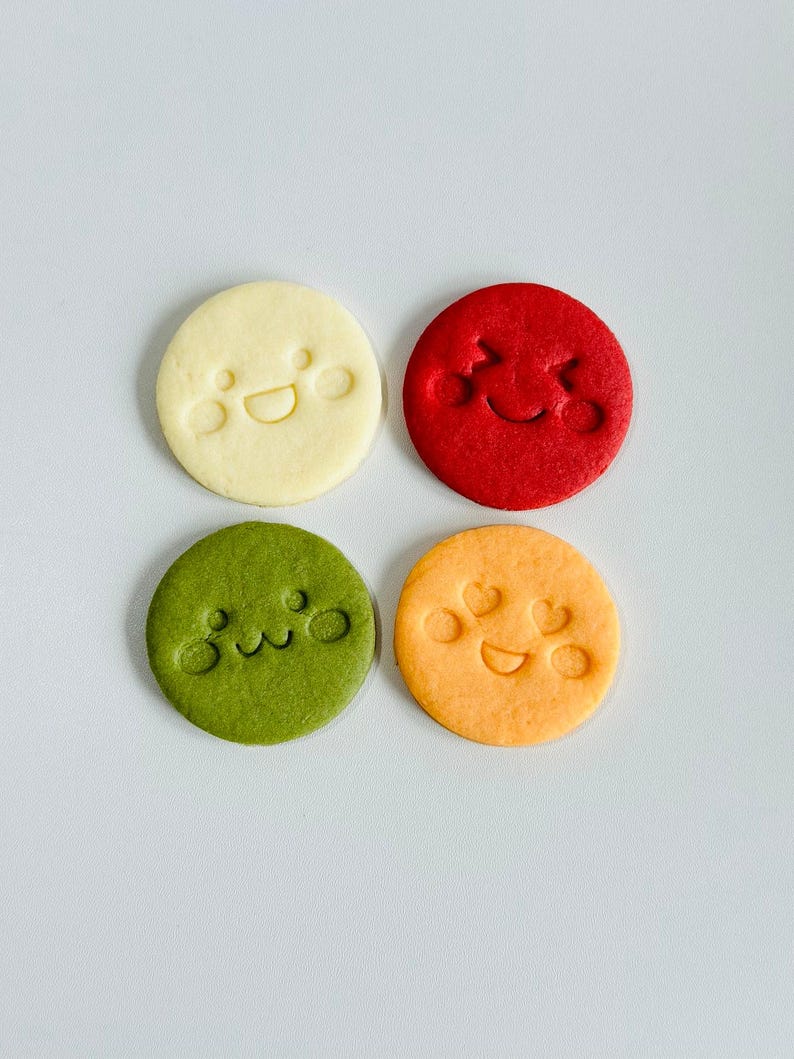 May include: Four round cookies in a square arrangement, each with a different color and a smiling face design. Colors include cream, red, green, and orange. The faces are created with simple, embossed lines.