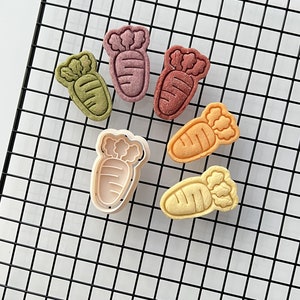 May include: Six carrot-shaped cookie cutters, including one with a white outline, on a black wire rack. The cutters are in various colors, including green, purple, orange, and yellow.