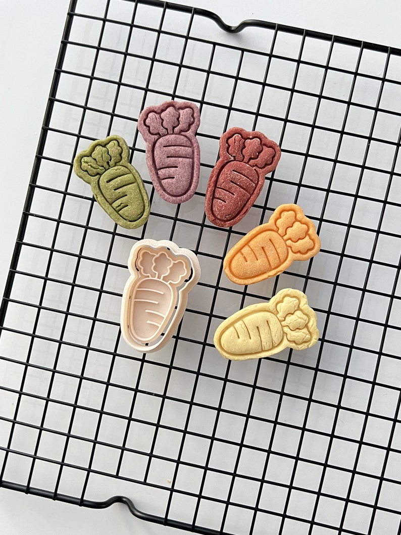 May include: Six carrot-shaped cookie cutters, including one with a white outline, on a black wire rack. The cutters are in various colors, including green, purple, orange, and yellow.