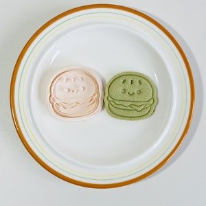 Puede incluir: Dos cortadores de galletas con forma de hamburguesa. Uno es rosa y el otro es verde. Ambos tienen una cara sonriente.