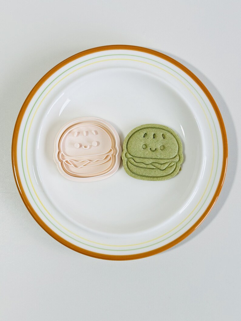 Puede incluir: Dos cortadores de galletas con forma de hamburguesa. Uno es rosa y el otro es verde. Ambos tienen una cara sonriente.