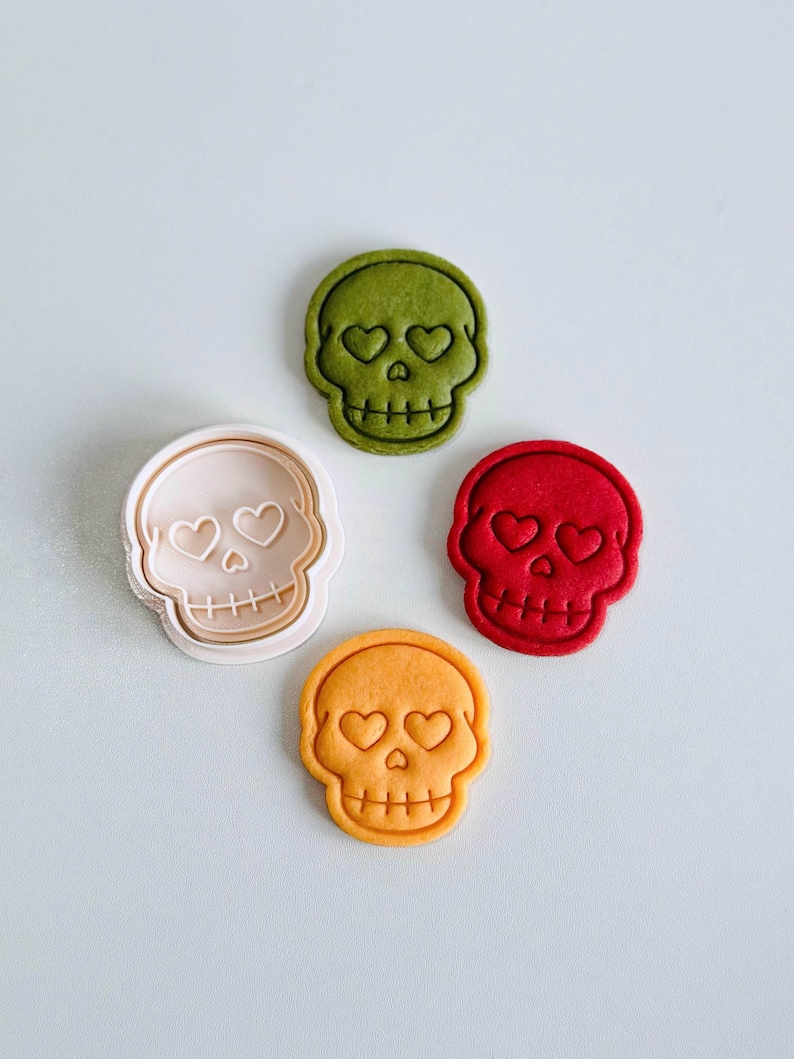 May include: Four skull-shaped cookie cutters and cookies. The cookies are in green, red, and orange, with heart-shaped eyes. A beige cookie cutter is also present. The cookies are on a white surface.