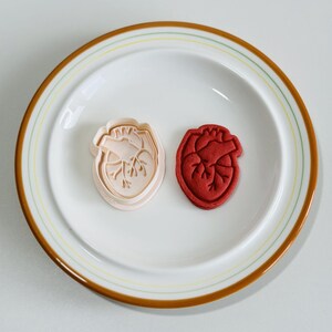 May include: Two heart-shaped cookie cutters, one pink and one red, on a white plate with a brown rim.