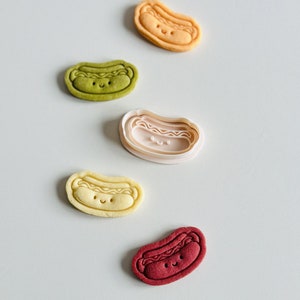 May include: Five different colored cookie cutters shaped like hot dogs with smiling faces. The cookie cutters are on a white surface.