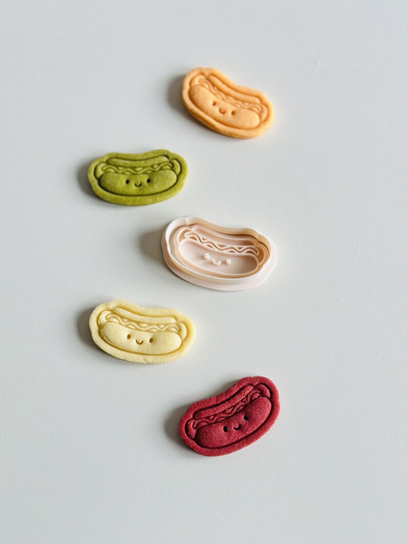 May include: Five different colored cookie cutters shaped like hot dogs with smiling faces. The cookie cutters are on a white surface.