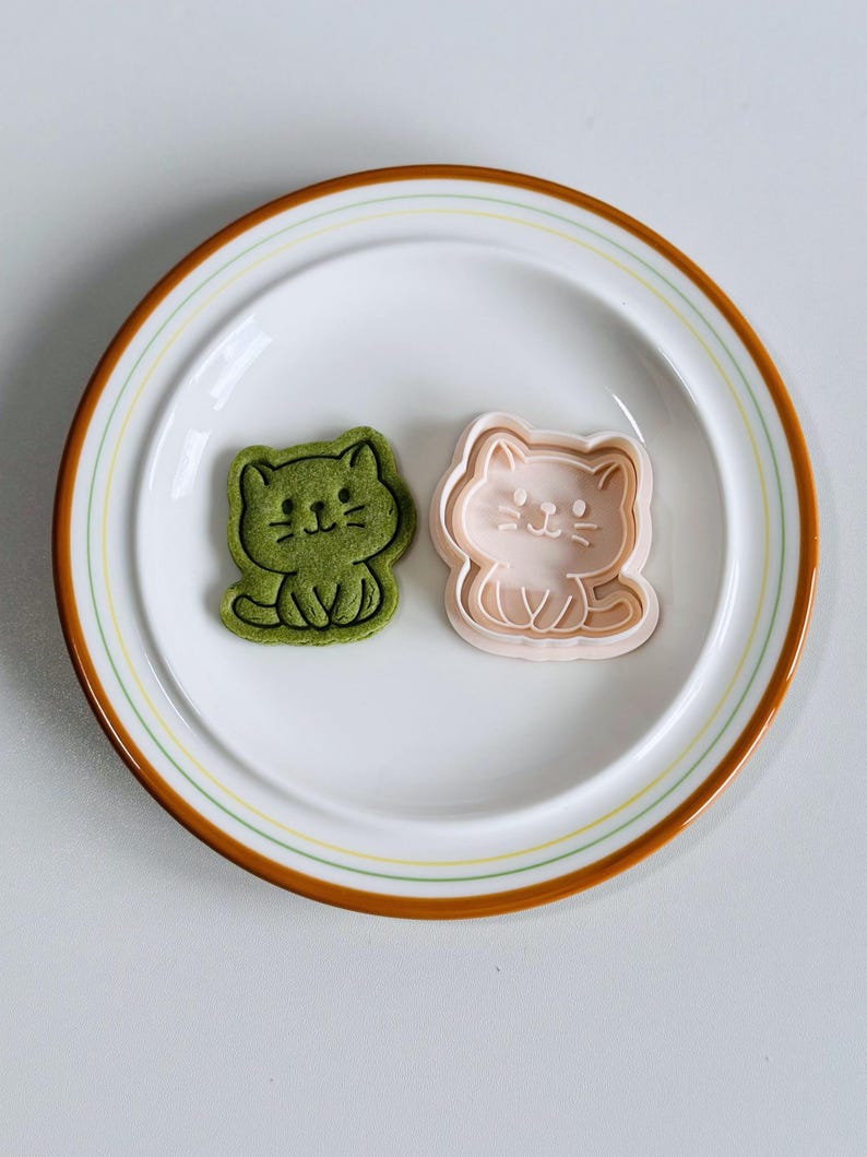 Puede incluir: Un plato blanco con borde marr&oacute;n y rayas verdes y amarillas contiene dos objetos con forma de gato. Uno es una galleta verde y el otro un cortador de galletas rosa. Los gatos est&aacute;n sentados con rasgos sencillos.
