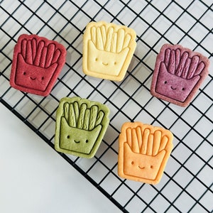 May include: Five decorated cookies shaped like a container of french fries. The cookies are yellow, orange, green, red, and purple. The cookies have a smiling face and are on a black wire rack.