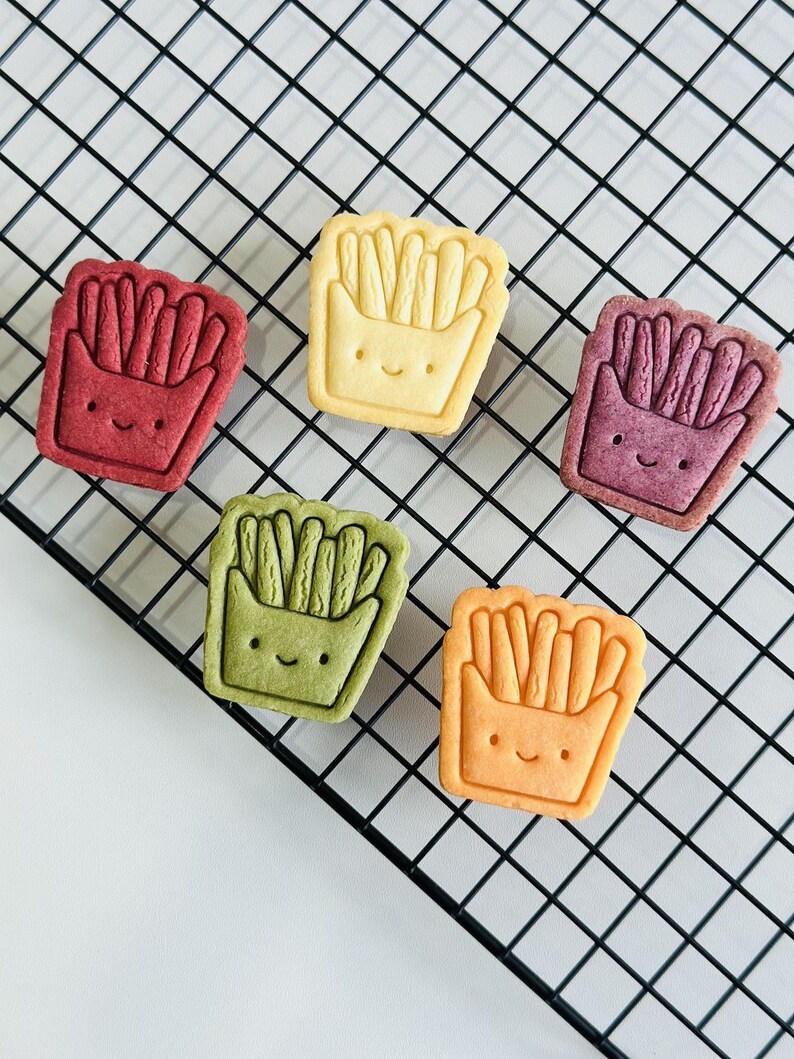 May include: Five decorated cookies shaped like a container of french fries. The cookies are yellow, orange, green, red, and purple. The cookies have a smiling face and are on a black wire rack.