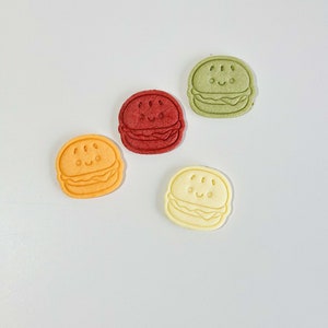 Puede incluir: Cuatro galletas de hamburguesa de estilo de dibujos animados de colores. Las galletas son en tonos de verde, rojo, naranja y amarillo.