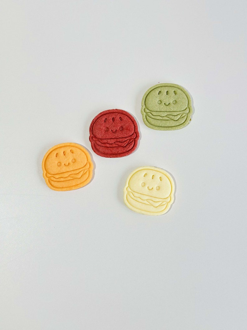 Puede incluir: Cuatro galletas de hamburguesa de estilo de dibujos animados de colores. Las galletas son en tonos de verde, rojo, naranja y amarillo.